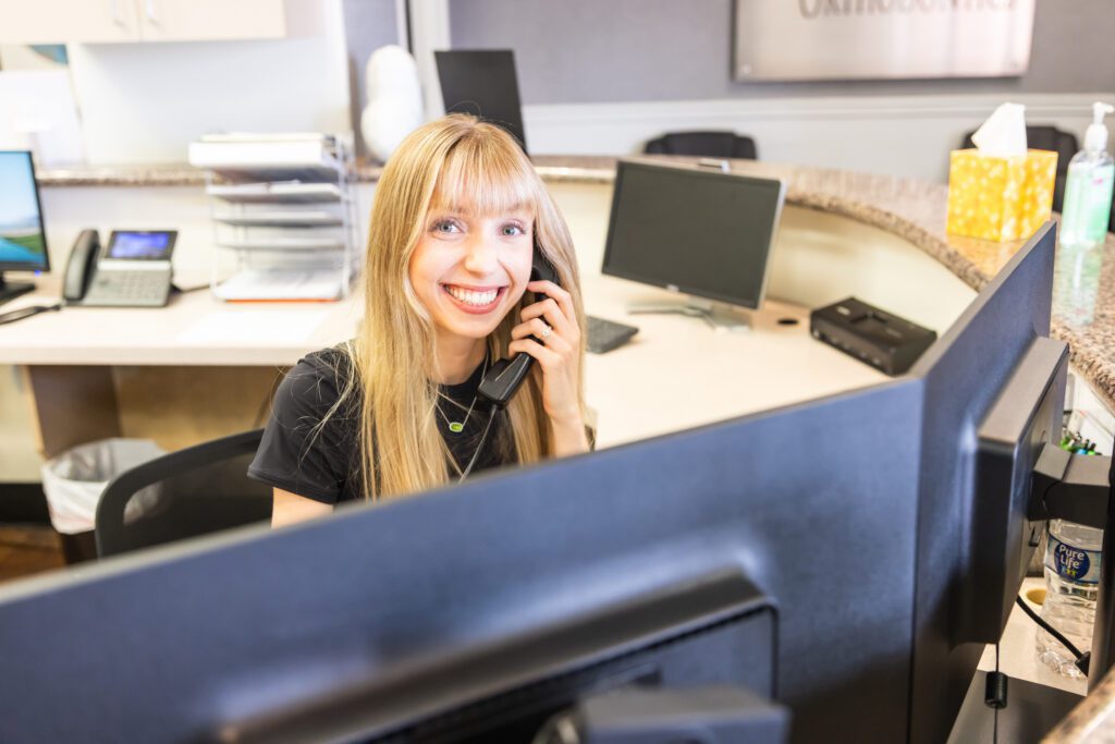 Receptionist smiling while answering a phone call at the front desk of a modern dental office.